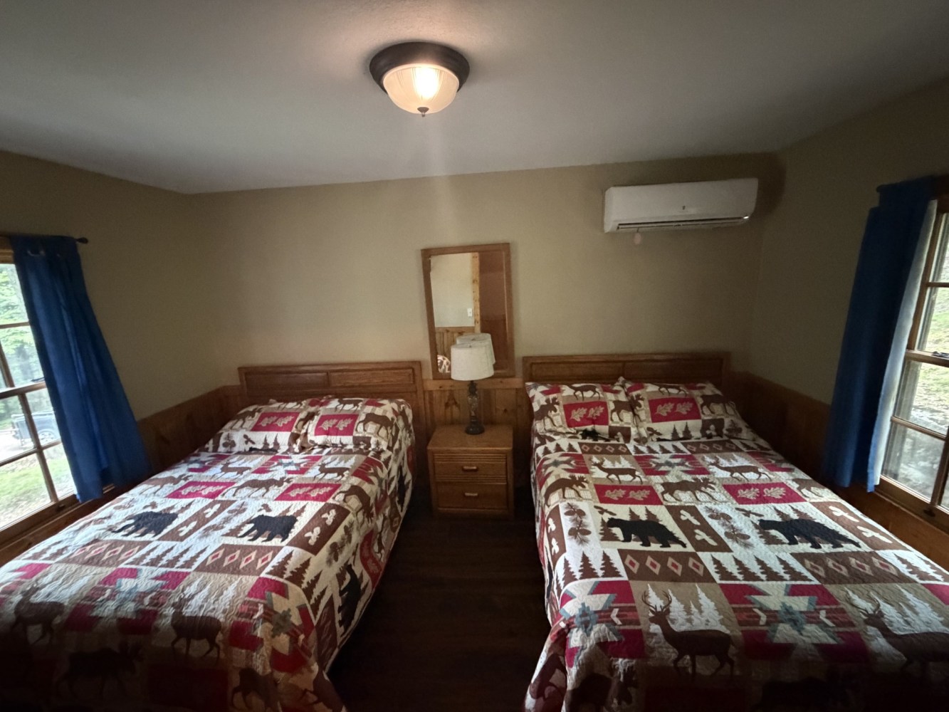 Room with two beds, colorful quilts, a shared nightstand, and windows with blue curtains.