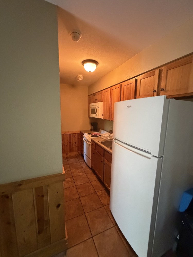 Narrow kitchen with wood cabinets, white appliances, and tiled floor.