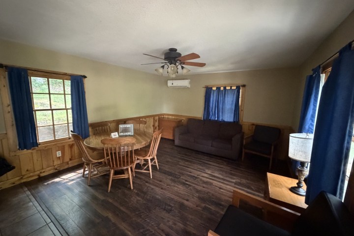 Cozy room with a table, chairs, sofa, lamp, and blue curtains, featuring wooden flooring and walls.