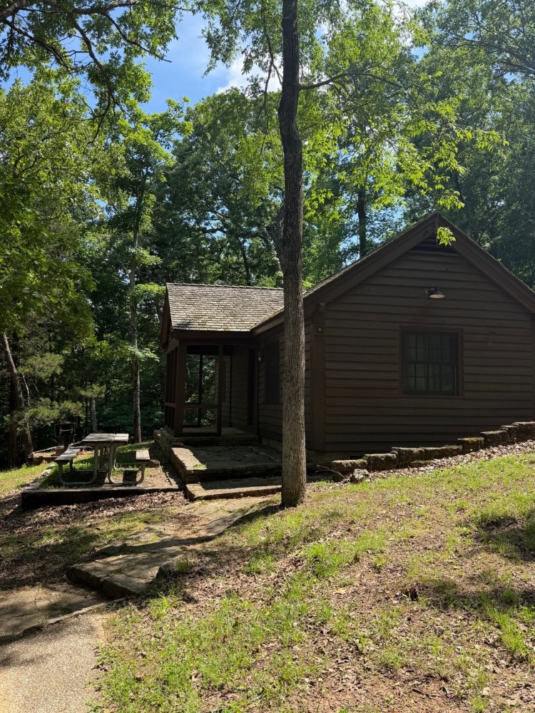 Small wooden cabin surrounded by trees with a picnic table outside.